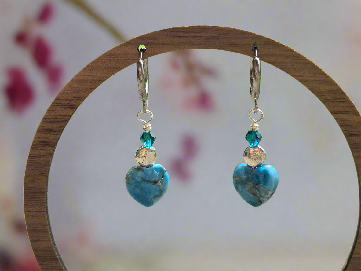 Blue heart-shaped earrings with silver and blue beads on a wooden stand with a blurred outdoor background.