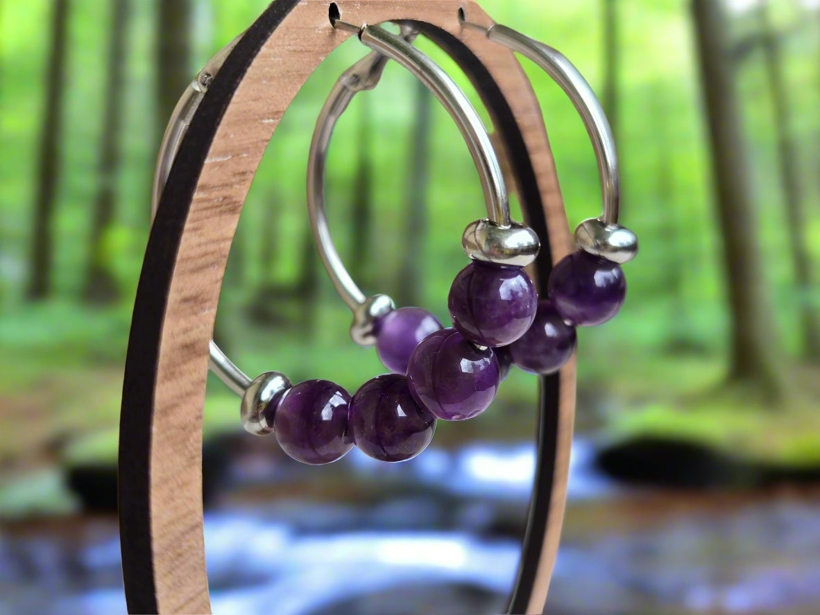Silver hoop earrings with purple beads on a wooden stand.